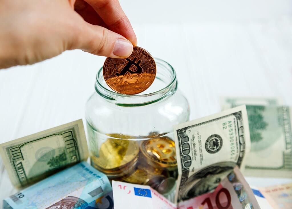 Womans hand puts a golden bitcoin into the glass jar among fiat money.