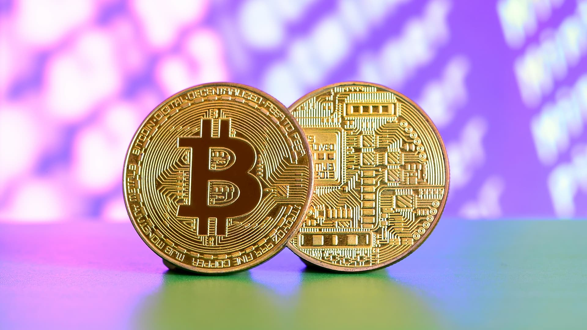 Two gold bitcoins lie on the green surface with a display in the background.