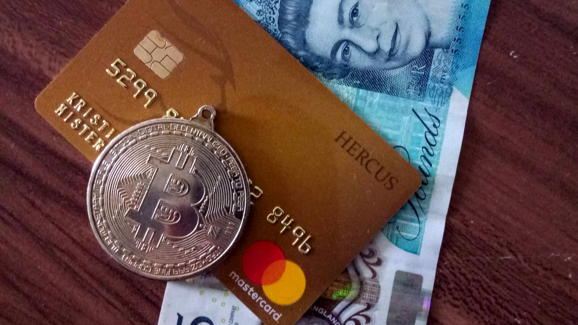 A five British pounds banknote with a golden plastic debit card Master Card and a metallic Bitcoin coin duplicate on a wooden surface. The representation of three generations of money: paper, electronic / plastic and digital.